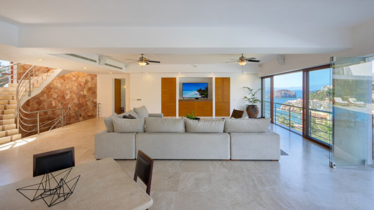 Villa Magnifico - Puerto Vallarta Luxury Rental A modern living room with a large white sectional sofa facing a flat-screen TV mounted on a wooden panel wall. To the right, glass doors offer views of Villa Magnifico Puerto Vallarta. A metal staircase with a stone accent wall leads upstairs, and a dining table with a geometric centerpiece is in the foreground.