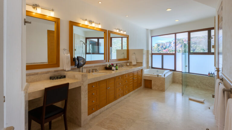 Villa Magnifico - Puerto Vallarta Luxury Rental Spacious modern bathroom at Villa Magnifico Puerto Vallarta featuring double sinks with large mirrors, a make-up station with a chair, a bathtub next to large windows with a scenic outdoor view, and a walk-in shower. The design includes beige stone countertops and wooden cabinetry.