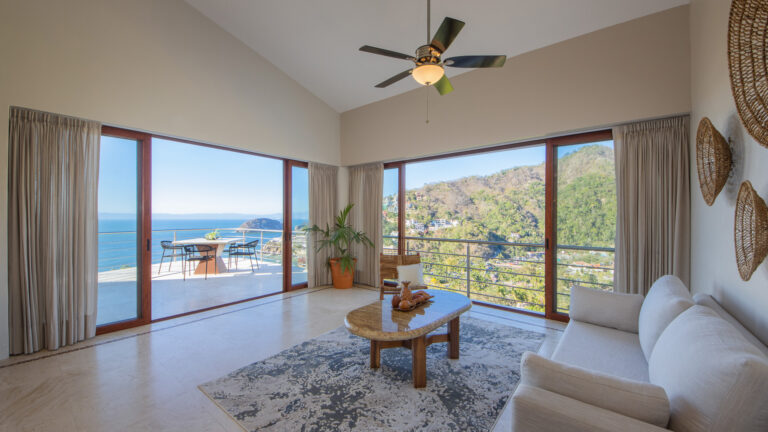 Villa Magnifico - Puerto Vallarta Luxury Rental The spacious, airy living room at Villa Magnifico Puerto Vallarta boasts floor-to-ceiling windows offering a panoramic view of the ocean and a green hillside. This elegant space features a beige sofa, wooden coffee table, ceiling fan, and patterned rug. Outside, enjoy the balcony with its dining table and chairs.