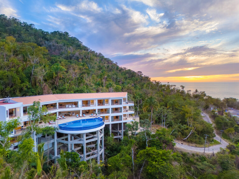 Villa Magnifico - Puerto Vallarta Luxury Rental Villa Magnifico Puerto Vallarta, a luxurious multi-story hotel with a rooftop infinity pool, is situated on a lush green hillside overlooking a scenic ocean view. The sky displays soft hues of pink, orange, and blue as the sun sets, creating a serene and picturesque tropical setting.