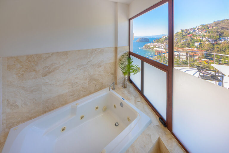 Villa Magnifico - Puerto Vallarta Luxury Rental A luxurious bathroom in Villa Magnifico Puerto Vallarta features a large Jacuzzi tub with beige marble tiles. The room offers a stunning ocean view through a large window lined with a thin palm leaf for a touch of greenery. Beyond the window are coastal cliffs and buildings nestled in the hills.