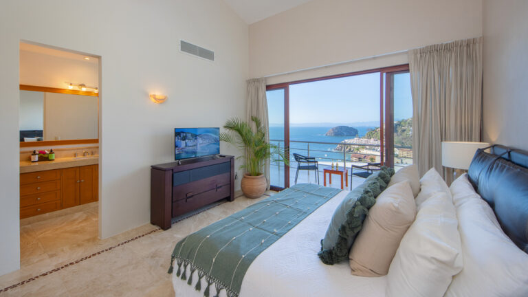 Villa Magnifico - Puerto Vallarta Luxury Rental A bright, airy bedroom in Villa Magnifico Puerto Vallarta features a king-sized bed, white linens, and green accents. The room offers a flat-screen TV, large dresser, potted plant, and en-suite bathroom. A glass door leads to a balcony with stunning ocean views of the coastline and lush greenery.