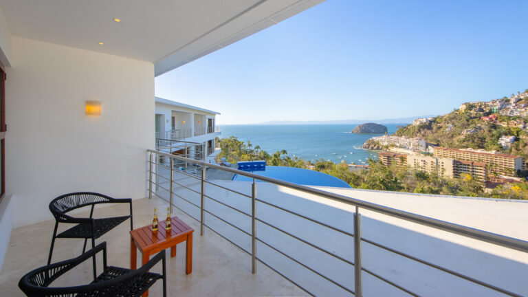 Villa Magnifico - Puerto Vallarta Luxury Rental A balcony view of the ocean and a distant island at Villa Magnifico Puerto Vallarta, with a white building to the left. Two wicker chairs and a small wooden table with two bottles are on the balcony. Below, buildings and greenery lead to the water. Clear blue sky completes the scenic vista.