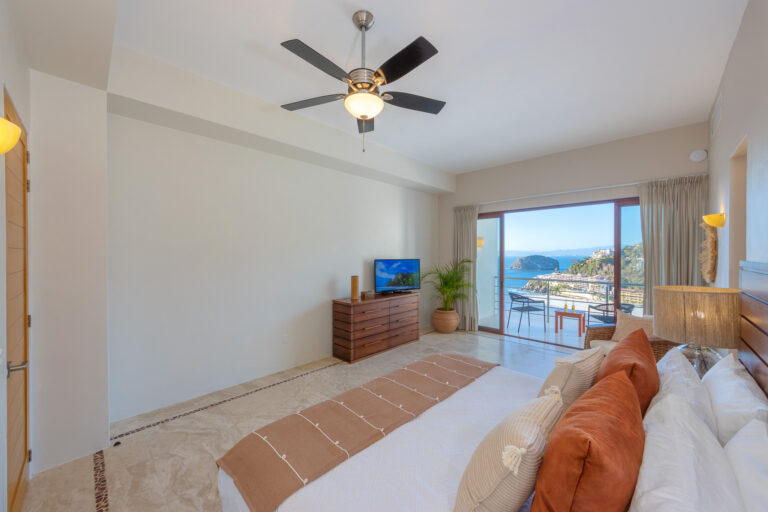 Villa Magnifico - Puerto Vallarta Luxury Rental A bright, modern bedroom in Villa Magnifico Puerto Vallarta features a double bed with orange and beige pillows, a ceiling fan, and a wooden dresser with a TV. Large glass doors provide a scenic view of the ocean and rocky coastline, opening to a spacious balcony with outdoor seating.