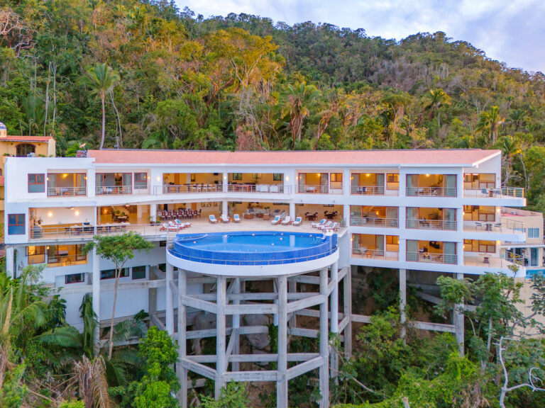 Villa Magnifico - Puerto Vallarta Luxury Rental Villa Magnifico Puerto Vallarta is a modern, multi-story hotel with a rooftop pool and large balconies, perched on a hillside surrounded by lush tropical vegetation. The building features white exteriors and expansive glass windows, offering stunning views of the forested landscape.