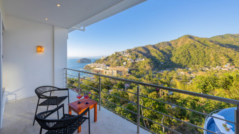 Villa Magnifico - Puerto Vallarta Luxury Rental A balcony at Villa Magnifico Puerto Vallarta with two black chairs and a small wooden table overlooks a scenic landscape with lush green hills, distant buildings, and the ocean meeting the clear blue sky. Two glasses with drinks are on the table, and warm light from a wall fixture glows softly.