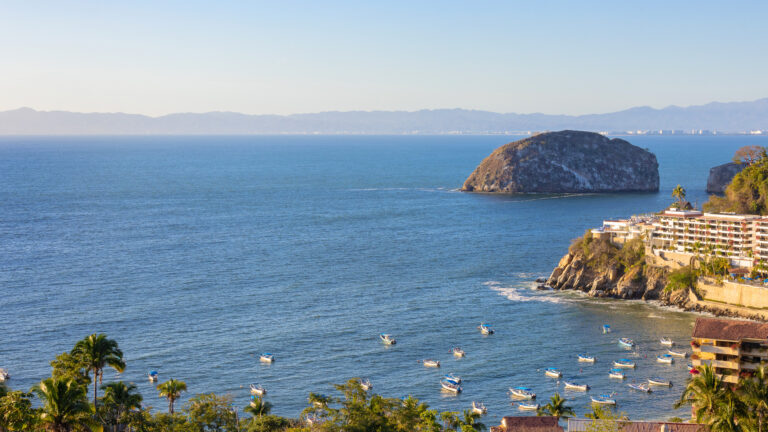 Villa Magnifico - Puerto Vallarta Luxury Rental A coastal scene with numerous small boats floating on calm blue waters. A large, rocky island is visible in the distance. There are buildings and palm trees on the shoreline, including the stunning Villa Magnifico Puerto Vallarta by Villa Experience, and the horizon features mountain ranges under a clear blue sky.