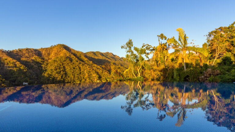 Villa Magnifico - Puerto Vallarta Luxury Rental A serene pool at Villa Magnifico Puerto Vallarta overlooks a lush, mountainous landscape under a clear blue sky. The water reflects the surrounding trees and mountains, blending the natural scenery with the tranquil surface of the pool. Tall trees can be seen scattered across the terrain.