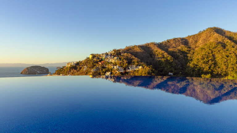 Villa Magnifico - Puerto Vallarta Luxury Rental An infinity pool at Villa Magnifico Puerto Vallarta by Villa Experience seamlessly merges with the horizon, reflecting blue skies and coastal hillside homes built among lush greenery. A small, rocky island is visible in the distance of the tranquil seascape under a clear sky.