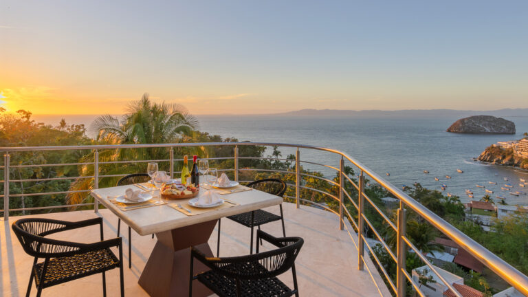 Villa Magnifico - Puerto Vallarta Luxury Rental A picturesque seaside dining setup at Villa Magnifico Puerto Vallarta boasts a table on a terrace overlooking the ocean at sunset. Elegantly set with glasses, plates, napkins, and a bottle of wine, palm trees and coastal rocks add to the scenic view, creating a serene, romantic ambiance.