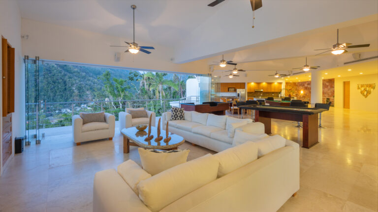 Villa Magnifico - Puerto Vallarta Luxury Rental A spacious, modern living room in Villa Magnifico Puerto Vallarta boasts large windows offering a scenic view of a lush, mountainous landscape. The room features white sofas, a coffee table with decorative candles, ceiling fans, a piano, a billiards table, and a kitchen with bar stools in the background.