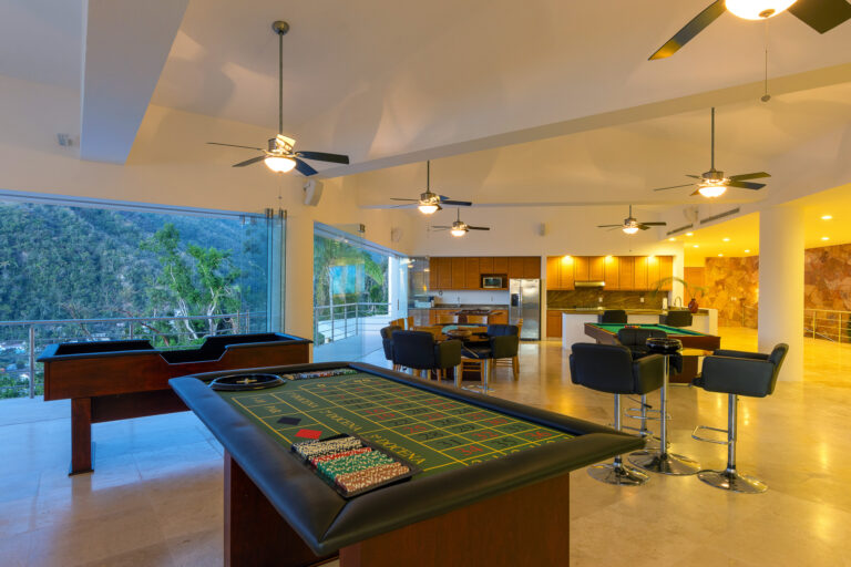 Villa Magnifico - Puerto Vallarta Luxury Rental A spacious, open-concept game room with a beautiful mountain view at Villa Magnifico Puerto Vallarta. The room features a poker table, a roulette table, a bar area with stools, and a foosball table. Large windows and ceiling fans add to the airy atmosphere, while the kitchen area is visible in the background.