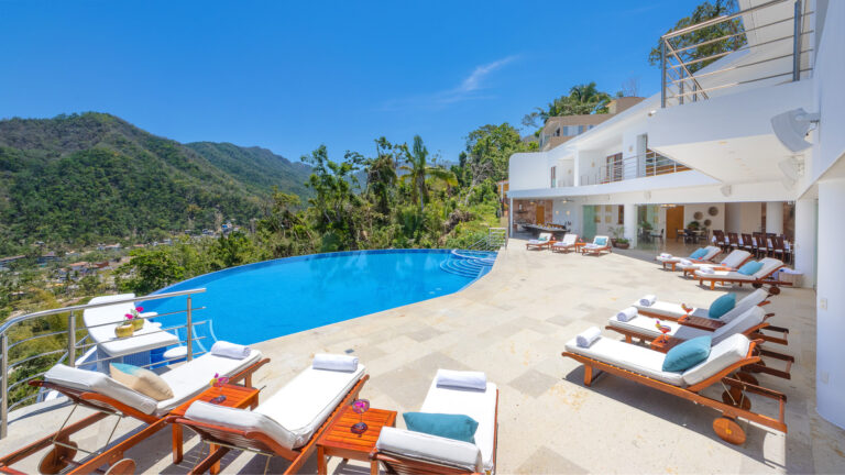 Villa Magnifico - Puerto Vallarta Luxury Rental The luxurious outdoor pool area at Villa Magnifico Puerto Vallarta features white lounge chairs and wooden tables set around an infinity pool overlooking lush green mountains and a clear blue sky. The modern white building has balconies and large windows, enhancing the scenic, tranquil ambiance.