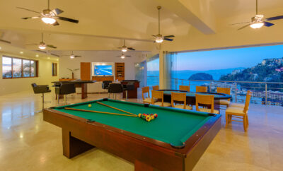 Villa Magnifico - Puerto Vallarta Luxury Rental A spacious, modern room at Villa Magnifico Puerto Vallarta boasts a pool table in the center, with floor-to-ceiling windows revealing a scenic coastal view. The room features ceiling fans, a dining area with wooden chairs and table, and a seating area with a wall-mounted TV and desk.
