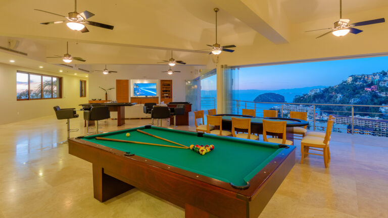 Villa Magnifico - Puerto Vallarta Luxury Rental A spacious, modern room at Villa Magnifico Puerto Vallarta boasts a pool table in the center, with floor-to-ceiling windows revealing a scenic coastal view. The room features ceiling fans, a dining area with wooden chairs and table, and a seating area with a wall-mounted TV and desk.