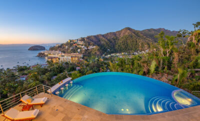 Villa Magnifico - Puerto Vallarta Luxury Rental An elevated view from Villa Magnifico in Puerto Vallarta showcases an infinity pool overlooking a coastal town with colorful buildings nestled between lush green hills. The pool features sun loungers on the deck, capturing a serene sunset over the ocean with a large rock formation near the shore.