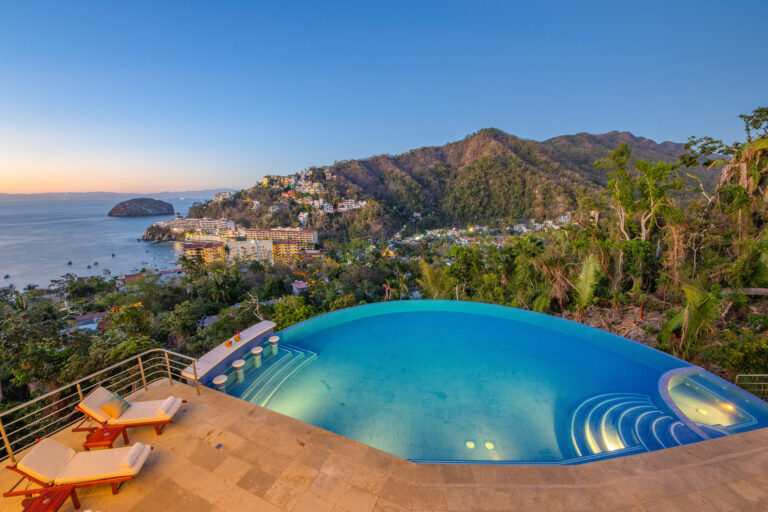 Villa Magnifico - Puerto Vallarta Luxury Rental An elevated view from Villa Magnifico in Puerto Vallarta showcases an infinity pool overlooking a coastal town with colorful buildings nestled between lush green hills. The pool features sun loungers on the deck, capturing a serene sunset over the ocean with a large rock formation near the shore.