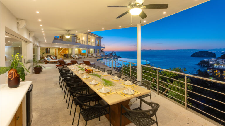 Villa Magnifico - Puerto Vallarta Luxury Rental The spacious outdoor dining area at Villa Magnifico Puerto Vallarta overlooks a stunning ocean view at sunset. The long table, set with plates, glasses, and napkins, is surrounded by black chairs. Ceiling fans and potted plants add comfort and greenery to the modern terrace.