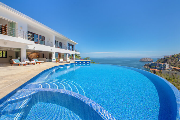 Villa Magnifico - Puerto Vallarta Luxury Rental Villa Magnifico in Puerto Vallarta offers a luxurious modern villa with a sleek design featuring an infinity pool overlooking the ocean. The patio has multiple lounging chairs and a seating area under a shaded porch, showcasing both the expansive pool and the panoramic view of the coastline and sky.