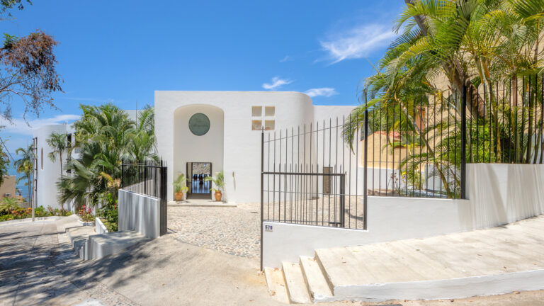 Villa Magnifico - Puerto Vallarta Luxury Rental A white modern building with smooth curved walls and a gated entryway surrounded by tall palm trees, Villa Magnifico in Puerto Vallarta boasts an arched doorway flanked by potted plants. Stairs and a pathway lead to the gate, and the sky is clear with a few clouds.