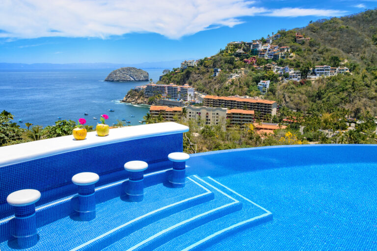 Villa Magnifico - Puerto Vallarta Luxury Rental An infinity pool with submerged bar stools at Villa Magnifico overlooks a coastal hill dotted with colorful buildings. The pool area features two yellow cocktails adorned with pink flowers. In the distance, a rocky outcrop juts into the blue sea under a partly cloudy sky in Puerto Vallarta.