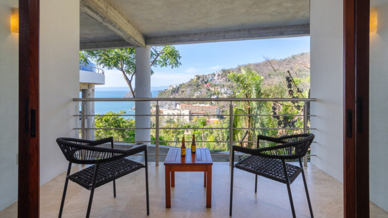 Villa Magnifico - Puerto Vallarta Luxury Rental A balcony at Villa Magnifico Puerto Vallarta features two black wicker chairs facing a small wooden table with two beer bottles. The balcony overlooks a picturesque coastal town with houses on a hillside and offers a clear view of the ocean, surrounded by lush greenery.