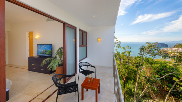 Villa Magnifico - Puerto Vallarta Luxury Rental A bright, open balcony at Villa Magnifico Puerto Vallarta features two black chairs and a small wooden table overlooking lush green foliage and a scenic ocean view. Adjacent is a room with a TV, dresser, potted plant, and an open door leading to another room. The sky is clear with a few clouds.