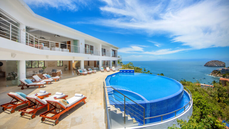 Villa Magnifico - Puerto Vallarta Luxury Rental Villa Magnifico Puerto Vallarta is a luxurious seaside villa with a spacious terrace featuring lounge chairs and a stunning infinity pool overlooking the ocean. The modern white building has large windows and balconies, offering panoramic views of the blue sky and expansive waters beyond.