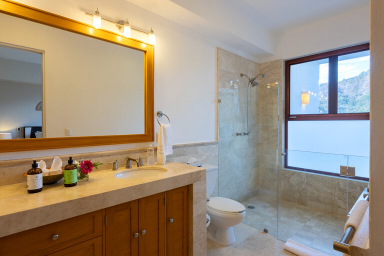 Villa Magnifico - Puerto Vallarta Luxury Rental A modern bathroom in Villa Magnifico Puerto Vallarta features a large mirror framed in wood above a stone countertop sink. The countertop holds two bottles and a small vase with pink flowers. A glass-enclosed shower is next to a toilet, while a window with natural light brightens the room.