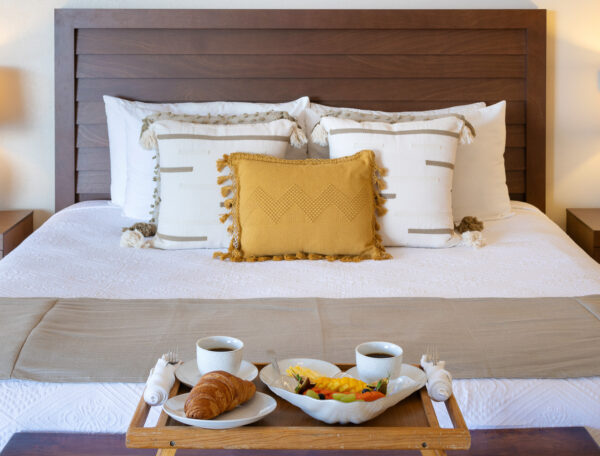 Villa Magnifico - Puerto Vallarta Luxury Rental A neatly made bed with white linens and pillows arranged against a wooden headboard in Villa Magnifico Puerto Vallarta. In front, a tray rests on the bed, holding two cups of coffee, a croissant, and a bowl of fresh fruit. Two bedside tables with lamps flank the bed.