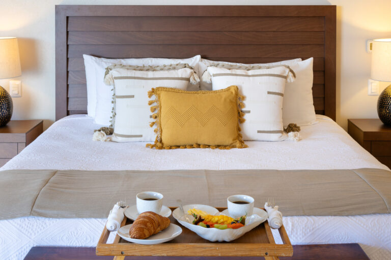 Villa Magnifico - Puerto Vallarta Luxury Rental A neatly made bed with white linens and pillows arranged against a wooden headboard in Villa Magnifico Puerto Vallarta. In front, a tray rests on the bed, holding two cups of coffee, a croissant, and a bowl of fresh fruit. Two bedside tables with lamps flank the bed.