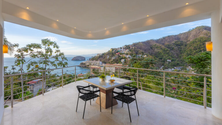 Villa Magnifico - Puerto Vallarta Luxury Rental A scenic balcony view at Villa Magnifico Puerto Vallarta overlooks a coastal landscape with hilly terrain, houses, and the ocean. The balcony features a dining table with chairs, and the area is lit by wall-mounted lights, creating a serene and inviting outdoor space.