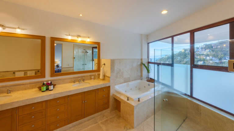 Villa Magnifico - Puerto Vallarta Luxury Rental A spacious bathroom with a large mirror above a double-sink vanity, ample cabinetry, a bathtub with jets, and a glass-enclosed shower. Large windows offer a view of the surrounding landscape and allow natural light to fill the room. Experience luxury at Villa Magnifico Puerto Vallarta.