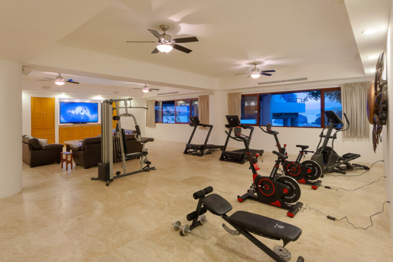 Villa Magnifico - Puerto Vallarta Luxury Rental A well-lit home gym at Villa Magnifico Puerto Vallarta features exercise equipment including treadmills, stationary bikes, weight machines, and benches. A sitting area with a TV and couches sits in the background. Large windows let in natural light, while ceiling fans are mounted on the white ceiling.