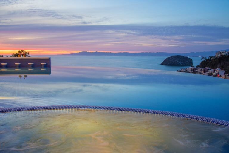 Villa Magnifico - Puerto Vallarta Luxury Rental A stunning view from the infinity pool at Villa Magnifico Puerto Vallarta overlooks a calm ocean at sunset. The sky is painted in hues of yellow, orange, and purple, with silhouettes of distant mountains and a large rock formation in the sea. The water reflects the vibrant colors of the sky.