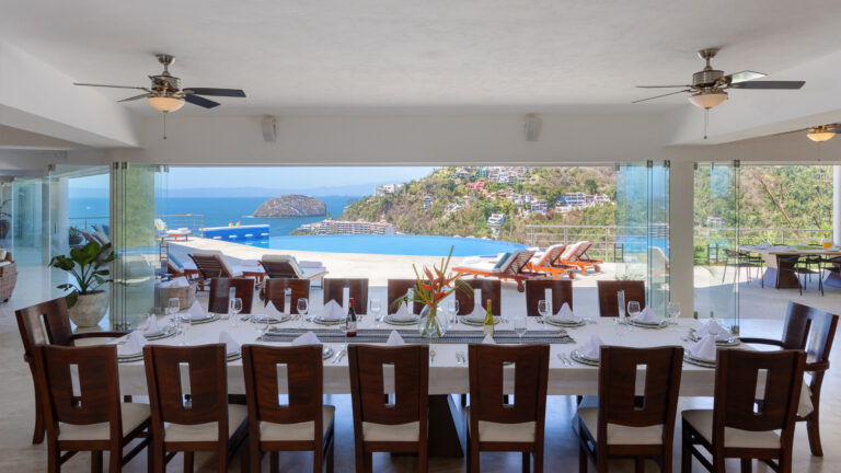 Villa Magnifico - Puerto Vallarta Luxury Rental A spacious dining room at Villa Magnifico in Puerto Vallarta boasts an elegantly set table for twelve, featuring wooden chairs and white tablecloths. Large glass doors open to a terrace with lounge chairs, showcasing a stunning ocean view and lush hillsides. A ceiling fan and potted plant add charm.