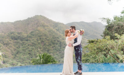 Villa Magnifico - Puerto Vallarta Luxury Rental A bride in a white lace gown and a groom in a white shirt with floral pants embrace each other by the poolside of Villa Magnifico, with lush, green mountains in the background. The bride holds a bouquet and they appear happy and in love. The atmosphere is serene and romantic.