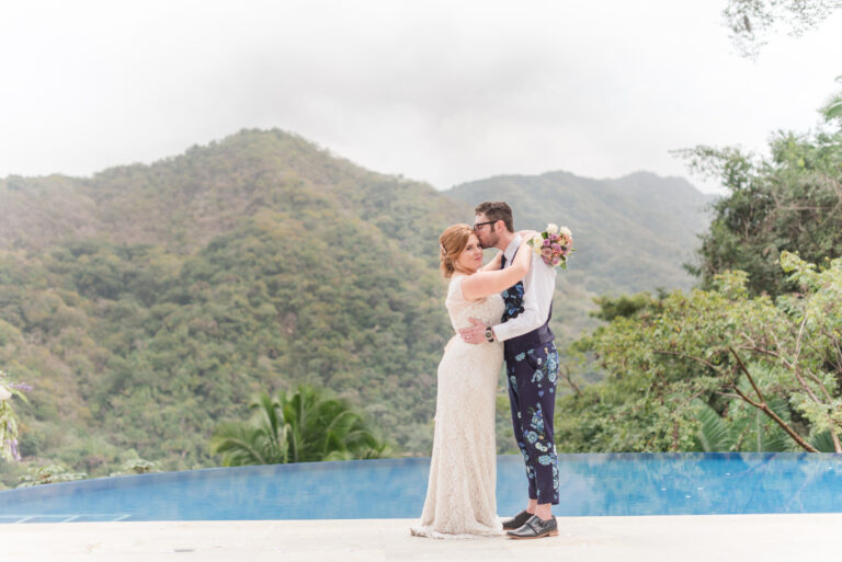 Villa Magnifico - Puerto Vallarta Luxury Rental A bride in a white lace gown and a groom in a white shirt with floral pants embrace each other by the poolside of Villa Magnifico, with lush, green mountains in the background. The bride holds a bouquet and they appear happy and in love. The atmosphere is serene and romantic.