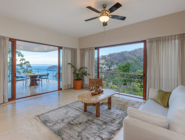 Villa Magnifico Luxury Villa in Mismaloya: Modern living room, white sofa, wooden table, ceiling fan; ocean-view balcony through glass doors.