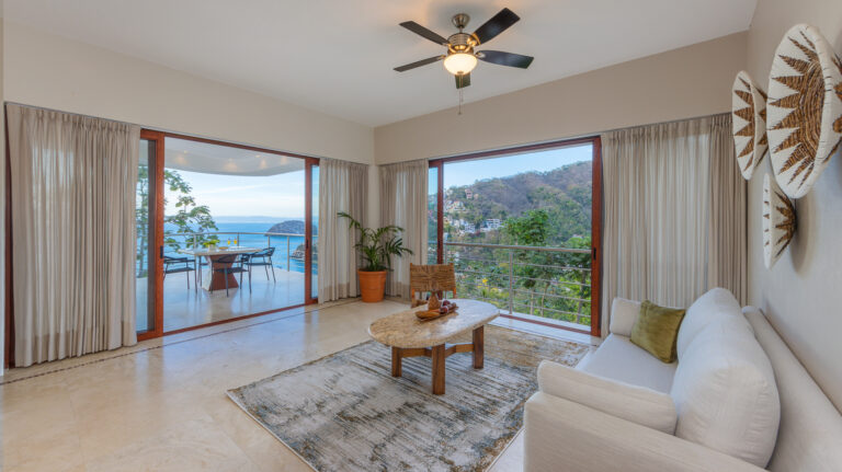 Villa Magnifico Luxury Villa in Mismaloya: Modern living room, white sofa, wooden table, ceiling fan; ocean-view balcony through glass doors.