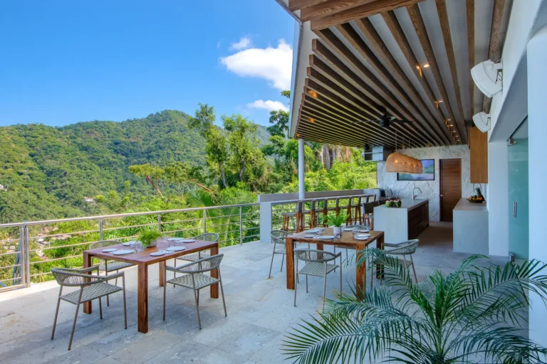 Villa Magnifico Outdoor dining area with wooden tables, open-air kitchen, lush green hills; in Mismaloya near Puerto Vallarta.