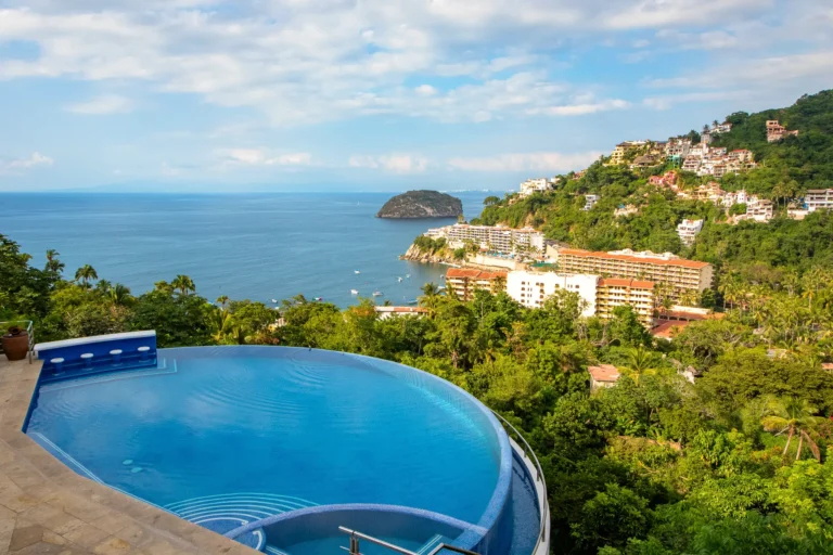 Villa Magnifico Luxury Villa in Mismaloya, near Puerto Vallarta: Infinity pool overlooking ocean, lush hills, and white buildings beneath a cloudy sky.