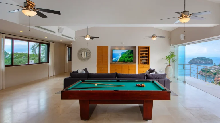 Villa Magnifico Luxury Villa in Mismaloya: Modern living room with pool table, ocean-view windows, and wall-mounted TV displaying a beach.