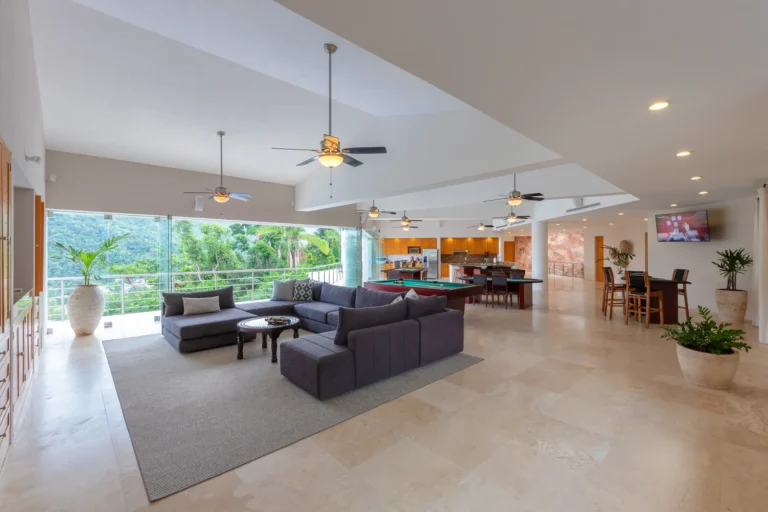 Villa Magnifico Modern living area with gray L-shaped sofa, large windows, ceiling fans; near Puerto Vallarta in Mismaloya.