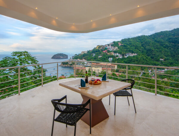 Villa Magnifico Balcony with table and chairs overlooks Mismaloya's ocean, lush hills, and a coastal town near Puerto Vallarta.