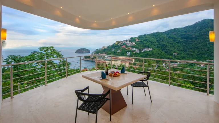 Villa Magnifico Terrace view in Mismaloya, near Puerto Vallarta: lush hillside, bay with island, shoreline buildings; table and chairs.