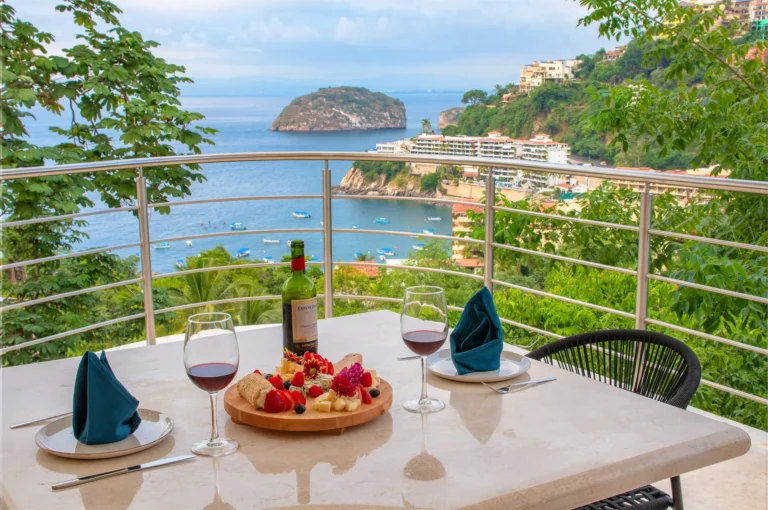 Villa Magnifico Ocean view from Mismaloya villa balcony; table with wine, glasses, napkins, cheese, fruit; hillside and island beyond.