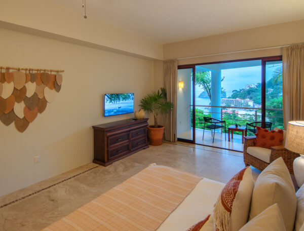 Villa Magnifico Hotel room with patterned bed, wooden dresser, leaf art; balcony overlooks greenery and ocean in Mismaloya, Mexico.