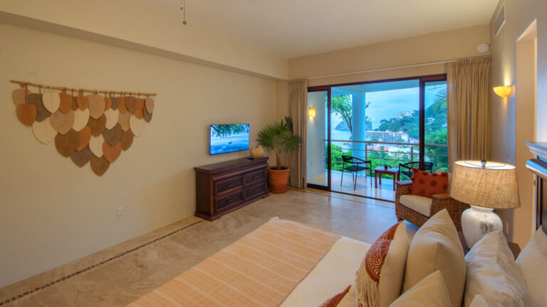 Villa Magnifico Hotel room with patterned bed, wooden dresser, leaf art; balcony overlooks greenery and ocean in Mismaloya, Mexico.
