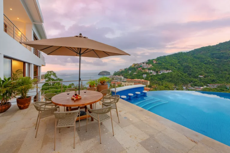 Villa Magnifico Luxury villa terrace in Mismaloya, Mexico: round table, chairs, umbrella; overlooks infinity pool, hillside, ocean sunset.